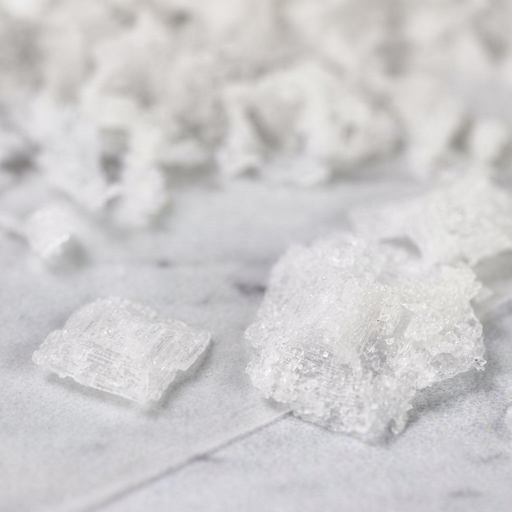 Close up of white crystalline Maldon salt flakes scattered on a light marble surface, with shallow depth of field focusing on the foreground pieces.