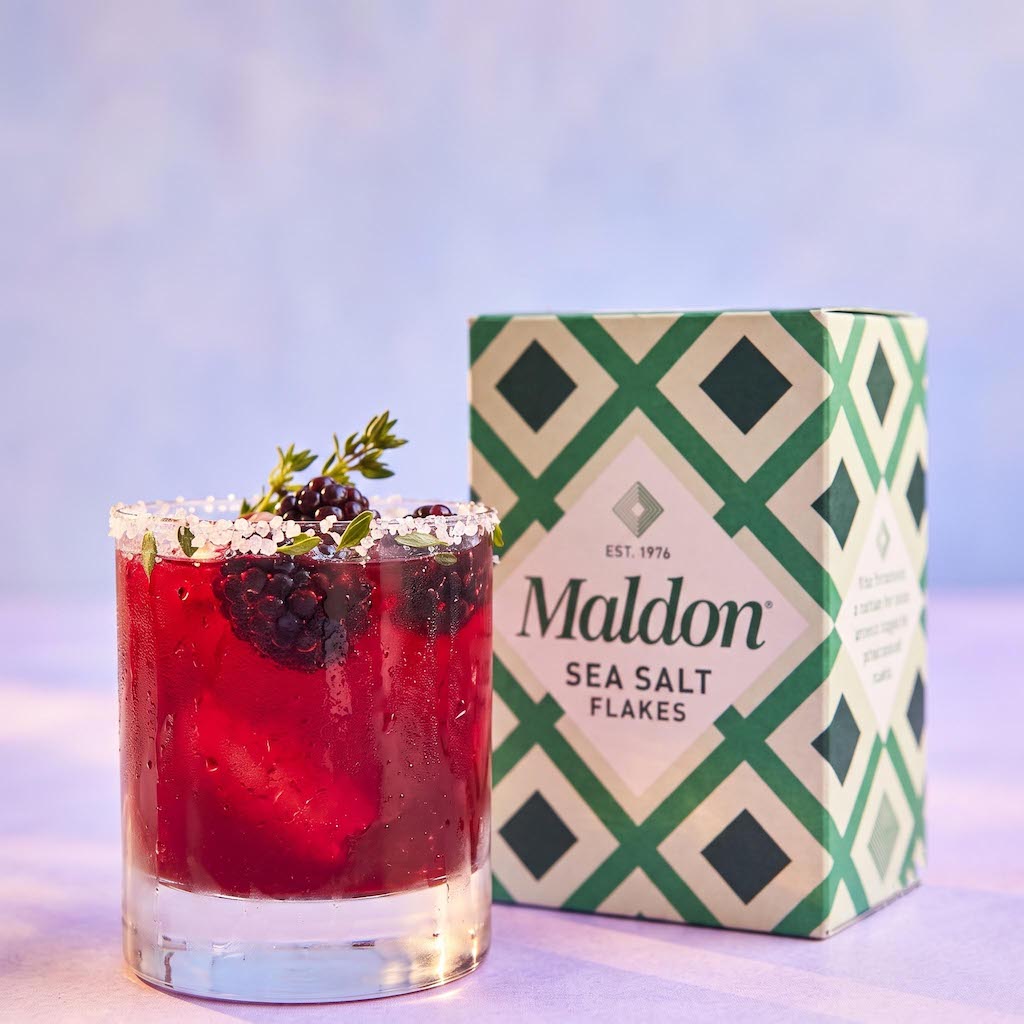 A glass of red cocktail with blackberries and a sprig of thyme sits beside a box of Maldon Sea Salt Flakes on a soft lavender surface. The box has a green and cream geometric pattern, and the glass has a Maldon white salt rim.