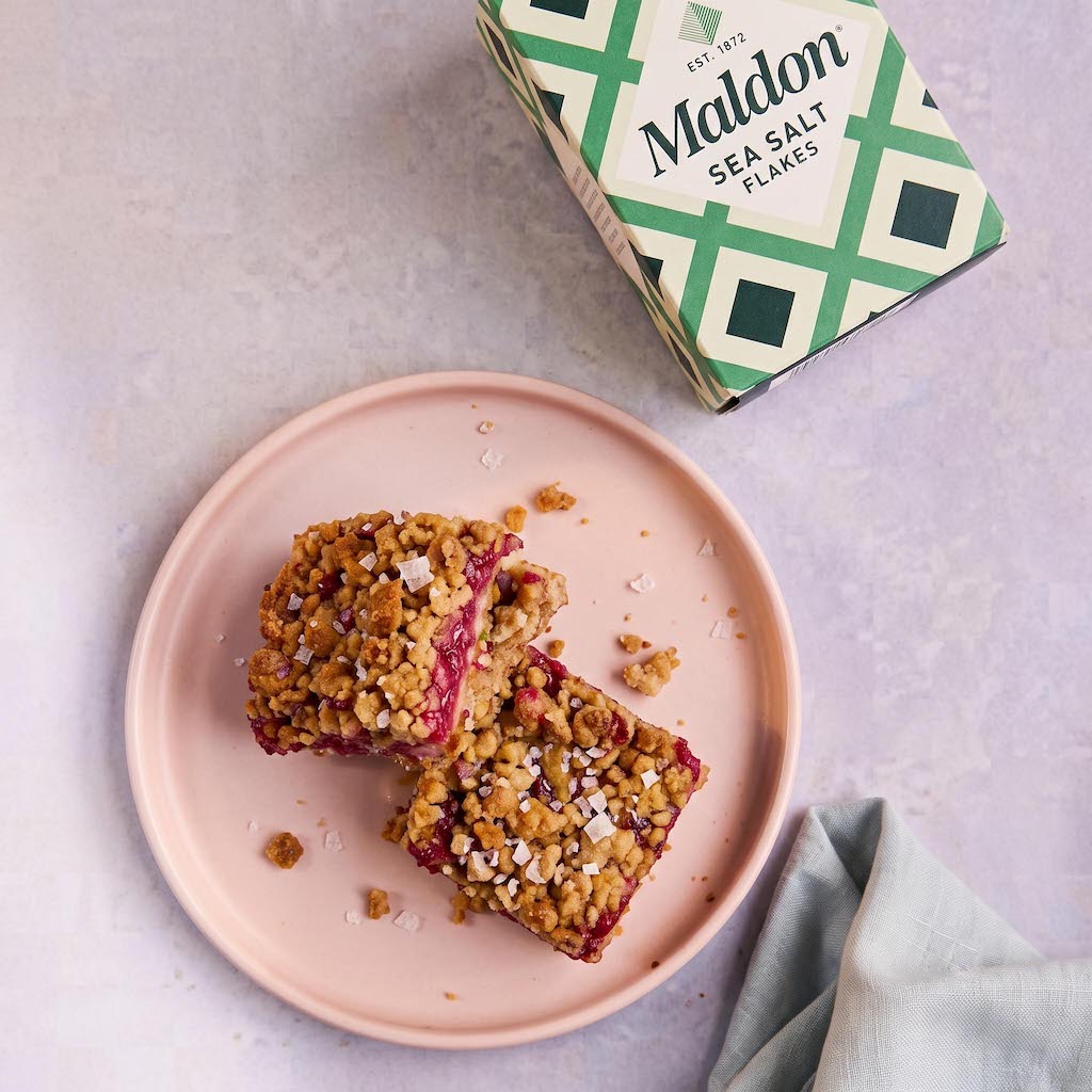 Two square sea salt flakes bars with a crumbly oat topping and red berry filling, served on a pink plate with the green and white Maldon Sea Salt Flakes box in the background.