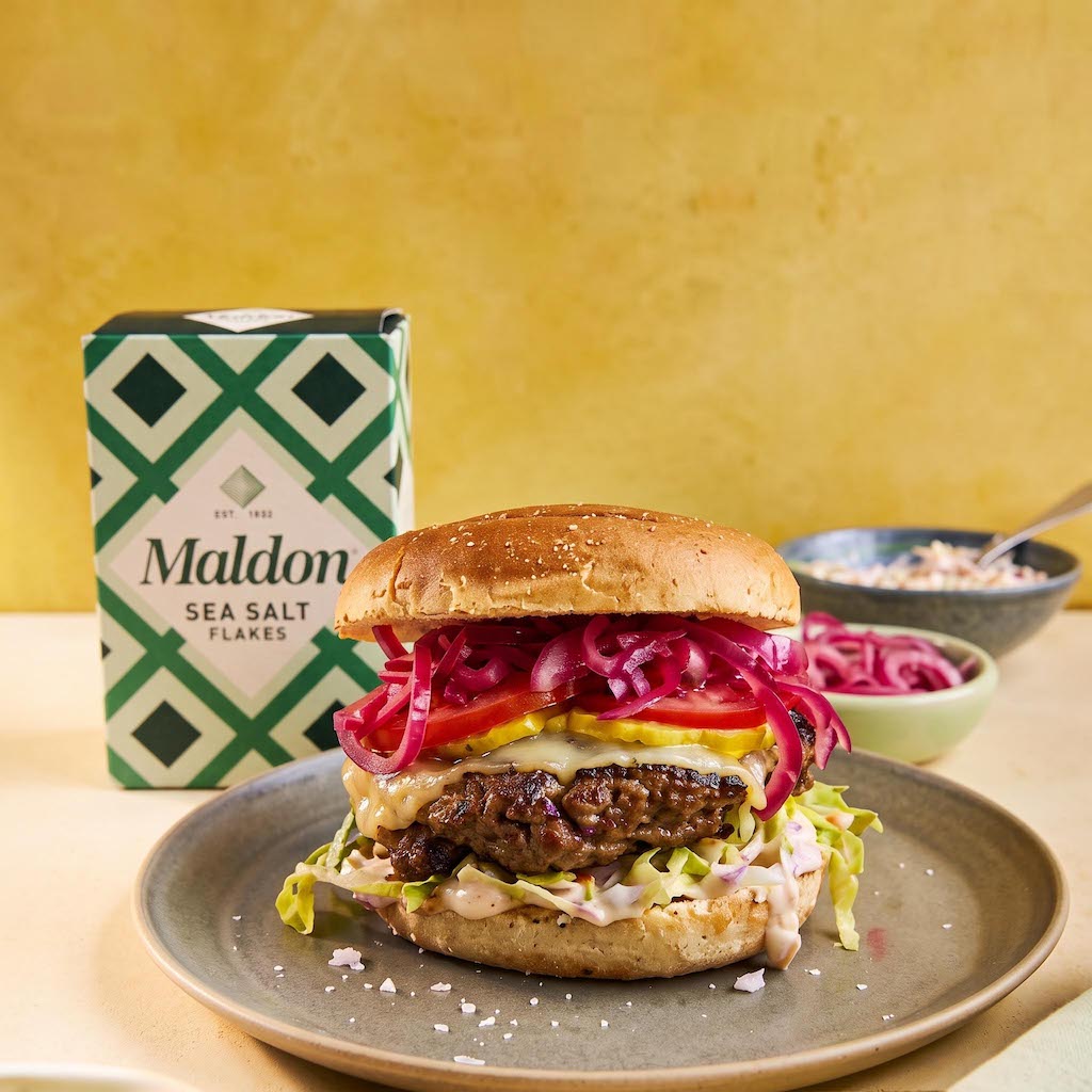 A gourmet burger with a beef patty, pickled red cabbage, tomato, and coleslaw on a plate, with a box of Maldon Sea Salt Flakes and bowls of garnish in the background against a textured yellow wall.