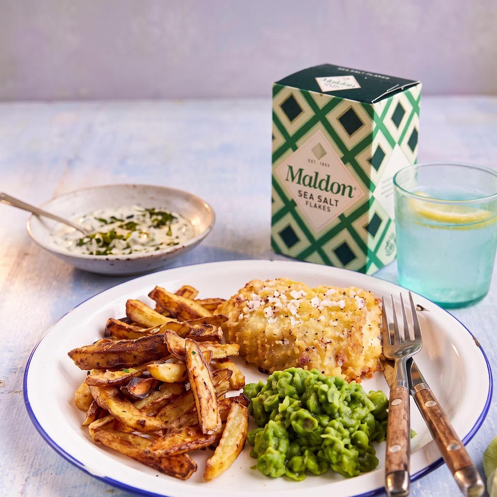 A plate of food with fries, mashed potatoes, and a baked item, styled with a glass of lemonade, a small bowl of dressing, and a box of Maldon Sea Salt Flakes in the background.