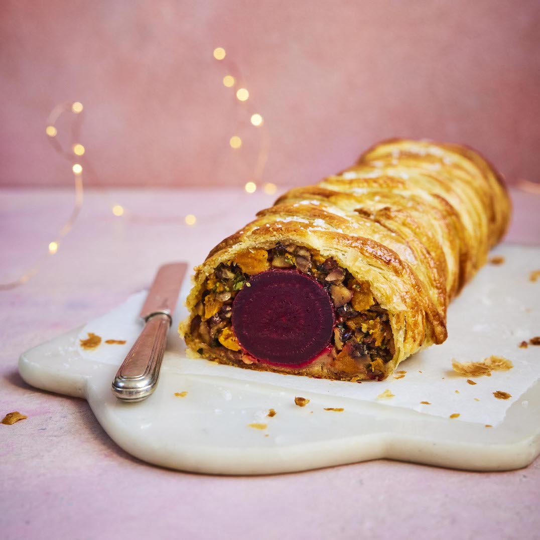 A baked pastry roll with a thick slice of red beet in the center, served on a white marble platter with a knife against a soft pink background with warm string lights.