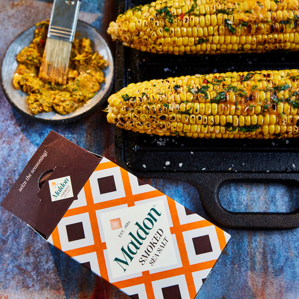 A package of Maldon smoked sea salt is shown in the foreground on a rustic blue surface, with two ears of grilled corn on a black tray and a small plate of buttered corn in the background.