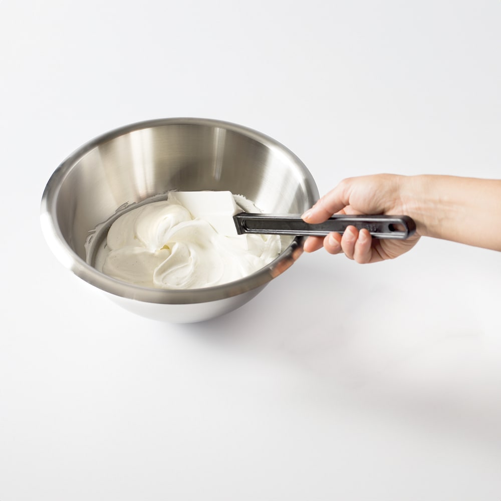 Person using a Matfer spatula to mix whipped cream in a metal bowl on a white background
