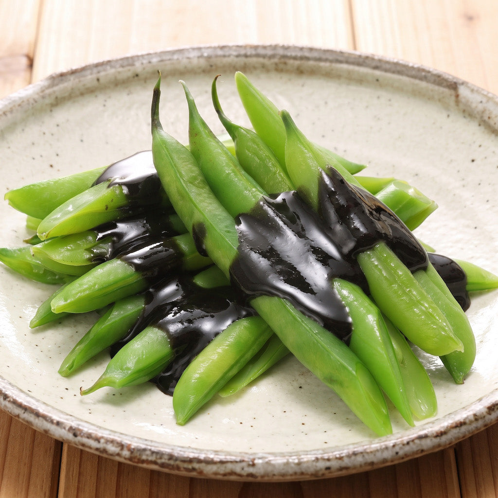 Stir-fried green beans with a glossy black sesame sauce, served on a speckled ceramic plate over a light wooden surface.