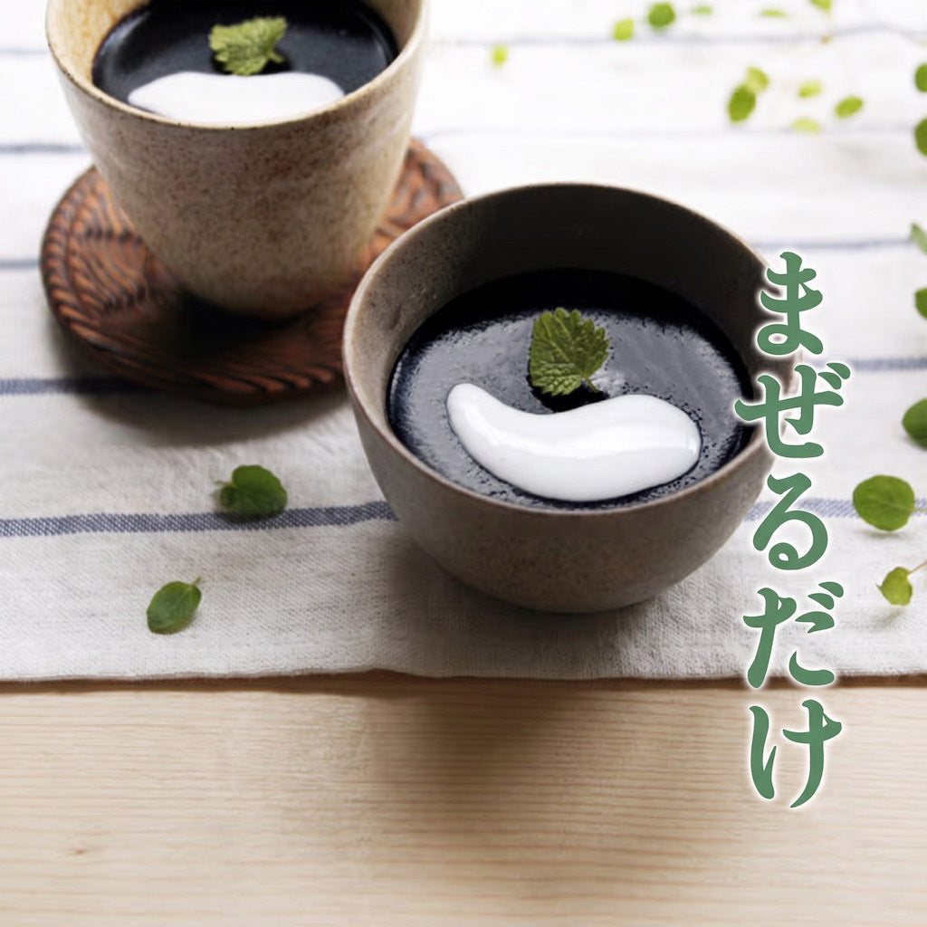 Two ceramic bowls containing a black sesame dessert or soup with a white swirl on top and a mint leaf, arranged on a striped cloth with Japanese text on the right side.
