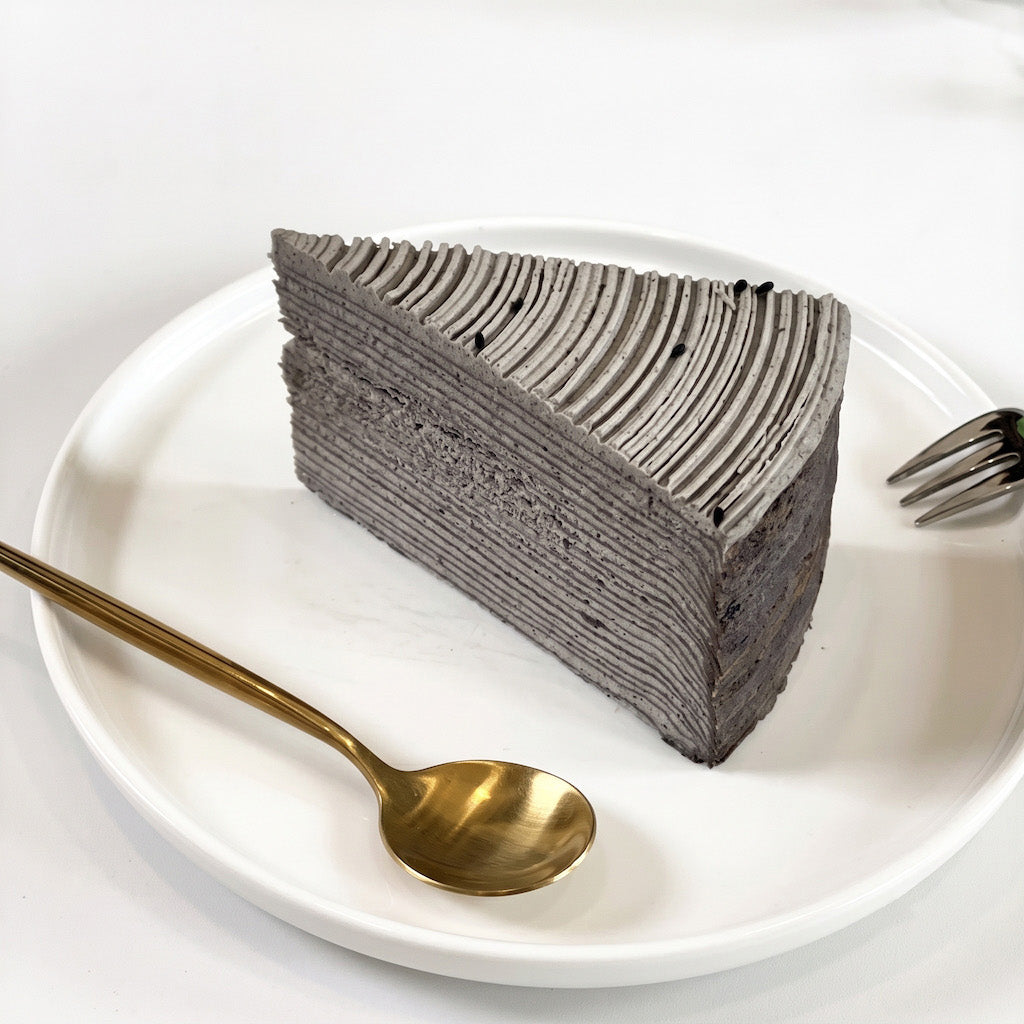 A triangular slice of layered black sesame crepe cake on a white plate, with a gold spoon and fork on a white surface.