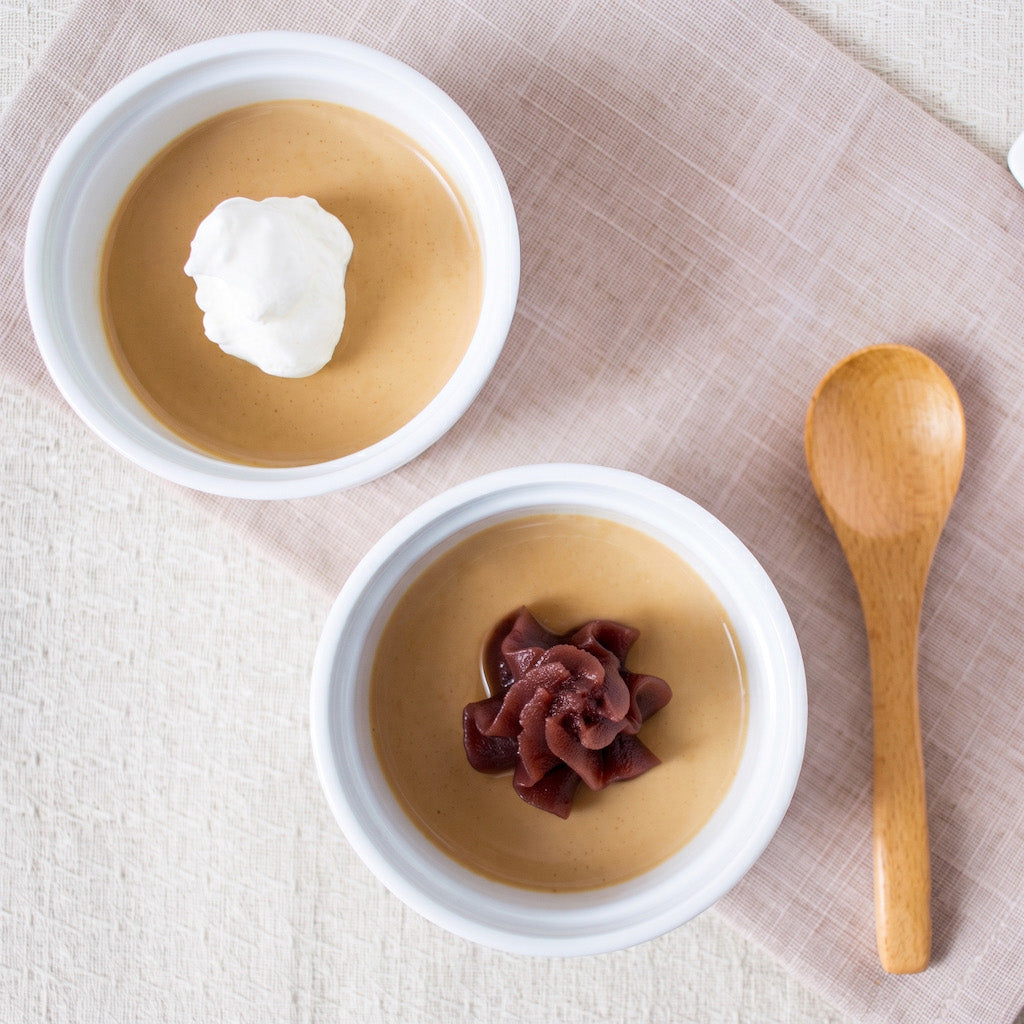 Two white bowls of custard dessert made with Kinako soy bean powder on a beige cloth, one topped with a dollop of whipped cream and the other with a dark berry garnish, with a wooden spoon on a textured beige surface.