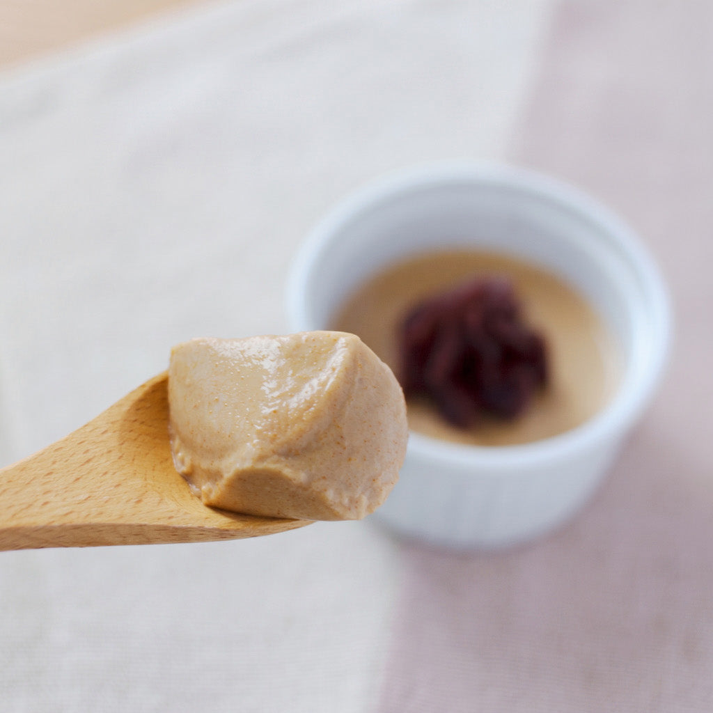 A close-up of a light brown creamy kinako dessert on a wooden spoon, with a small white bowl blurred in the background containing a dark red topping, set on a pale pink surface.