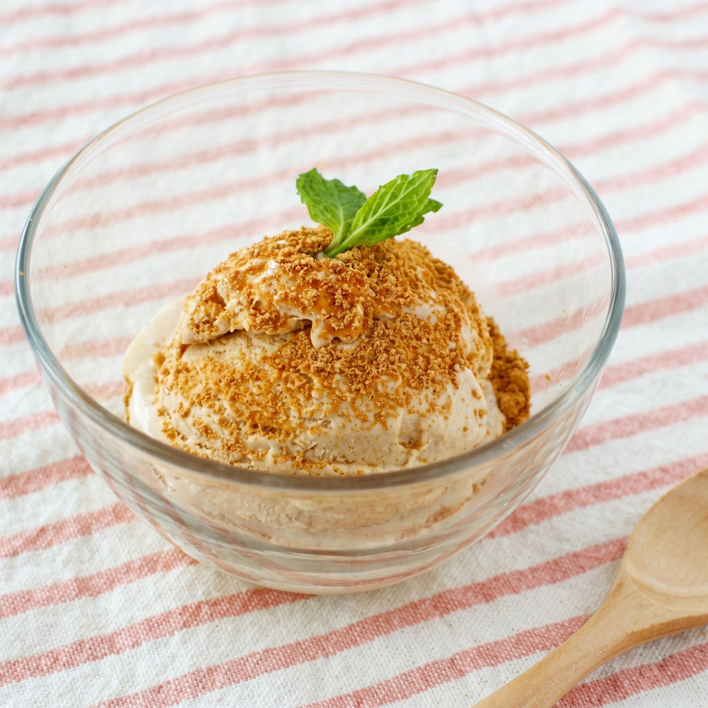 A scoop of light brown kinako roasted soy bean ice cream in a clear glass bowl, topped with a green mint leaf and a sprinkle of golden-brown crumbs, set on a pink and white striped cloth with a wooden spoon partially visible.