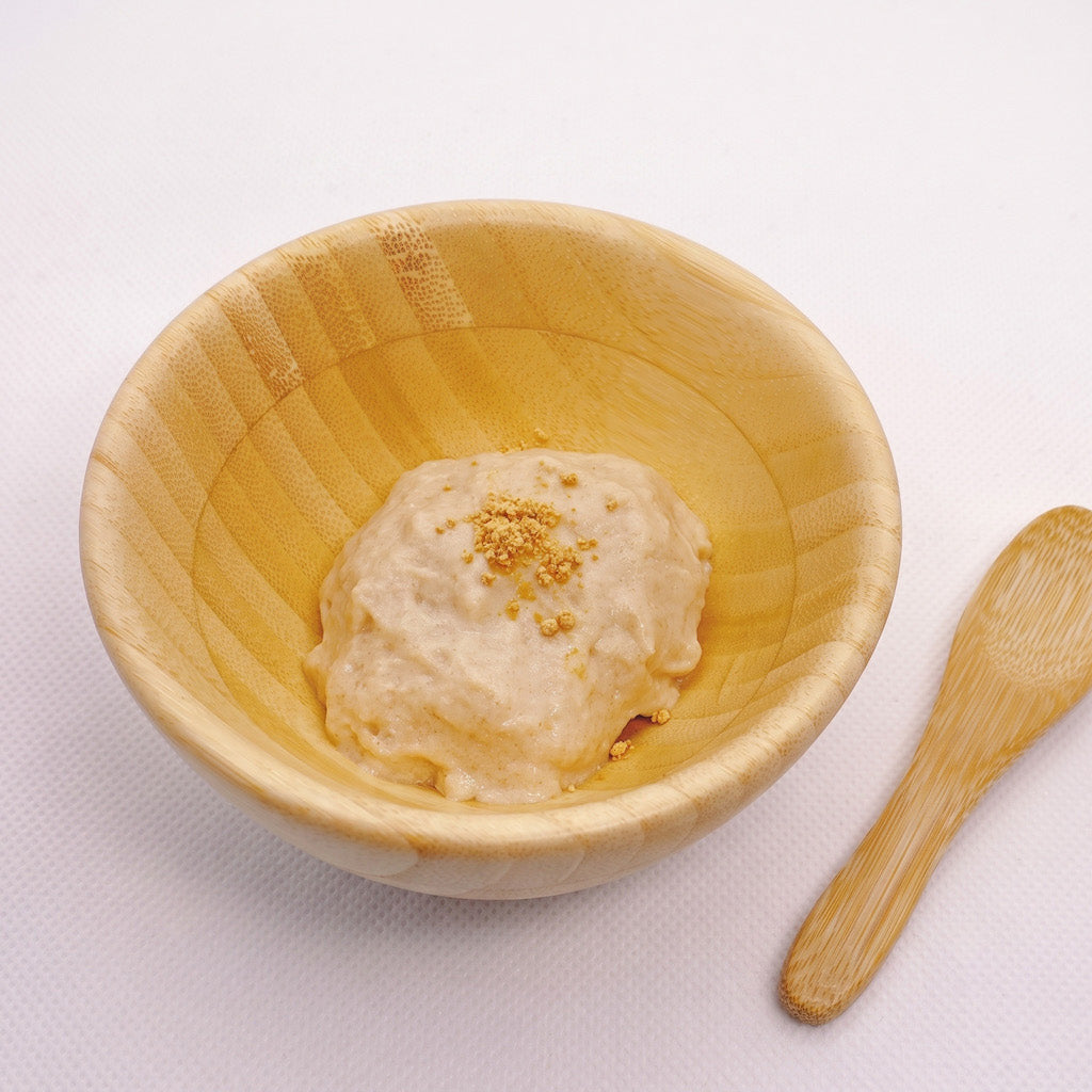 A light natural wood bowl containing a scoop of beige kinako dessert with a small wooden spoon placed to the right on a white textured background.