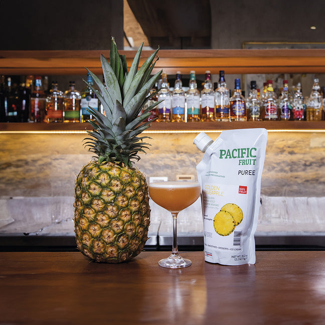 Pineapple next to a bag of Pacific Fruit Puree and a cocktail in a bar setting.