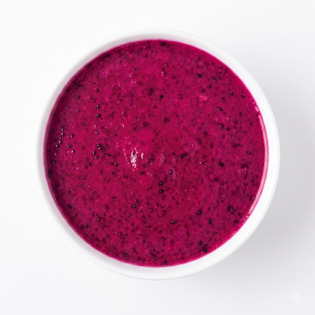 A top-down view of a white bowl filled with bright magenta-red dragon fruit puree, containing small dark seeds, on a clean white background.