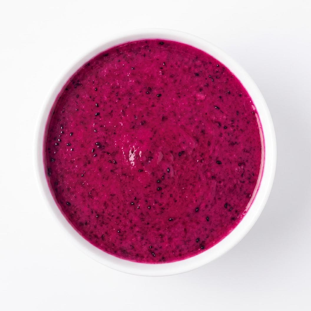 A top-down view of a white bowl filled with bright magenta-red dragon fruit puree, speckled with small black seeds, on a clean white background.