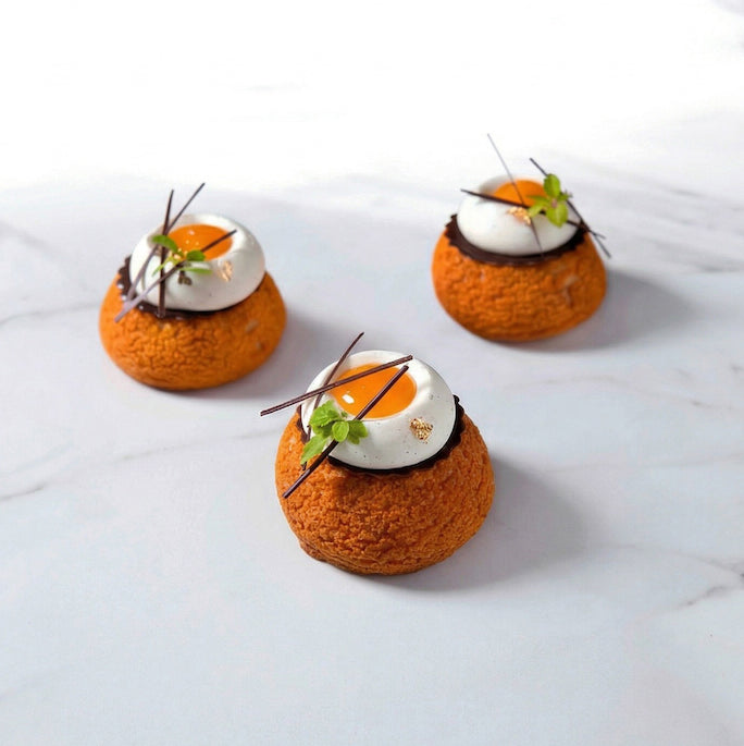 Three small dessert items with orange puff bases, white cream with a turmeric puree centre, and green garnishes on a light surface.