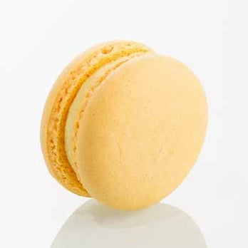 A single pale yellow macaron with a smooth, slightly glossy shell and light cream-colored filling, standing upright on a reflective white surface against a white background.