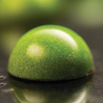 A close-up of a glossy green dome-shaped chocolate, resting on a dark reflective surface with a blurred green background.