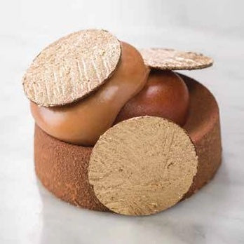 A close-up of a small round brown dessert or pastry with a smooth caramel-colored top and several textured round biscuit-like pieces placed on top and beside it, set on a light marble surface.