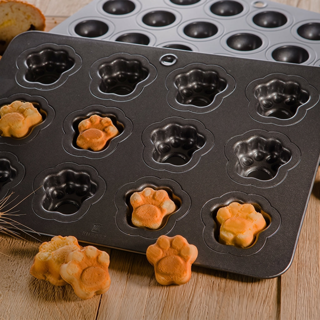 A dark gray non-stick Sanneng baking pan with paw-shaped cavities, shown with several baked golden-brown paw-shaped cookies or biscuits placed in and around the pan on a wooden surface.