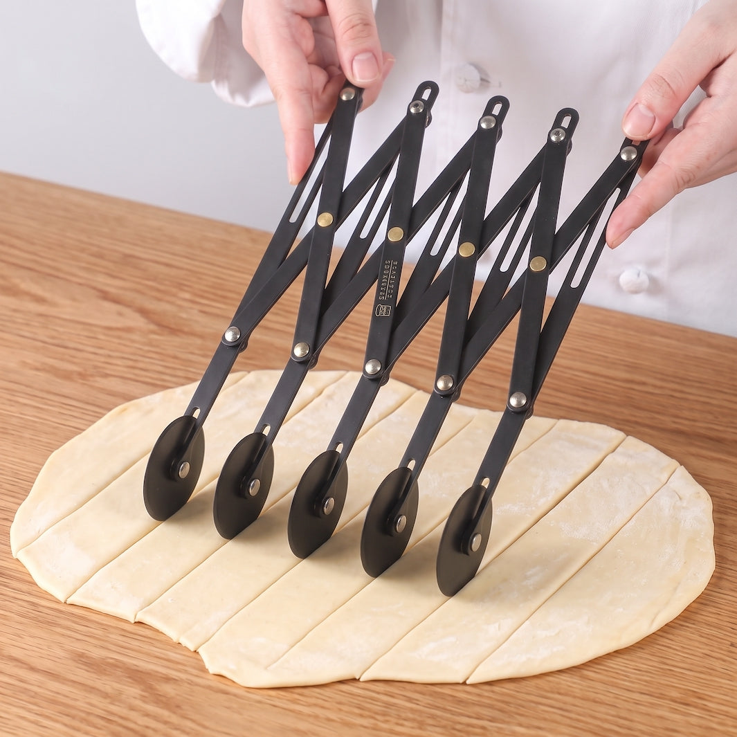 A set of Sanneng five black collapsible dough cutters with metal wheels and silver joints, being held by hands over a round piece of dough on a wooden table.
