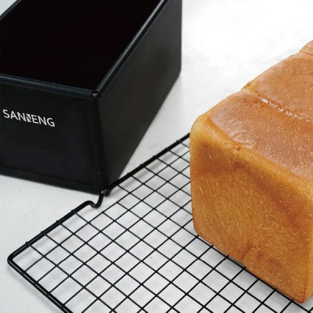 Loaf of bread on a cooling rack next to a black Sanneng Aluminized Steel loaf pan with 'SANNENG' branding.