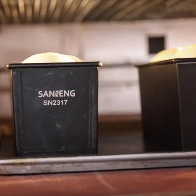 Two black aluminized steel loaf pans with 'SANNENG' branding on a blurred background