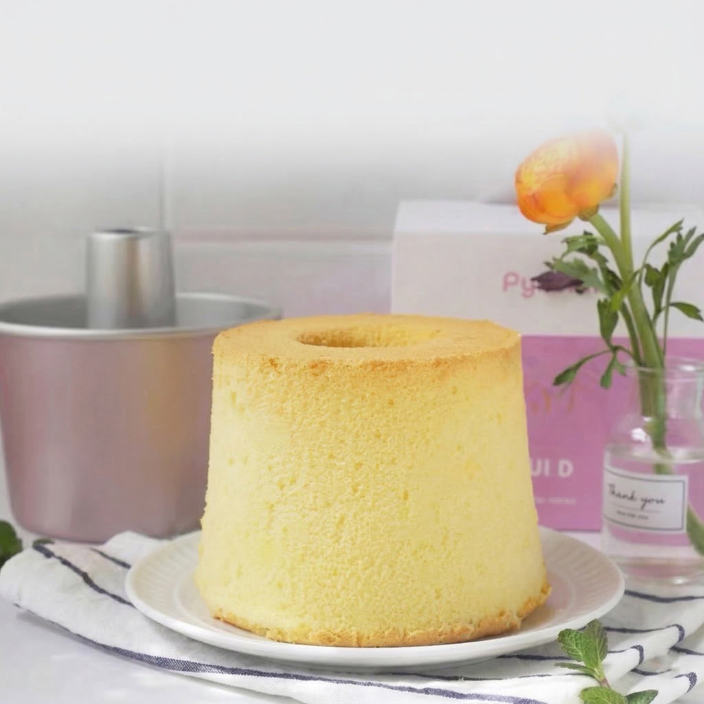 Yellow chiffon cake on a white plate with a pink box and flower vase in the background
