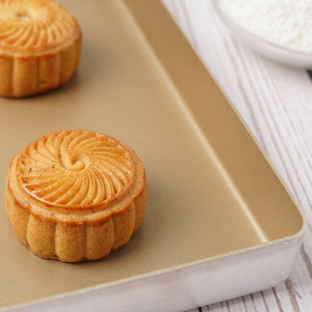 Two golden-brown mooncakes with a swirled top pattern resting on a rectangular gold Sanneng baking pan, set on a white wooden surface with a small bowl of white powder blurred in the background.