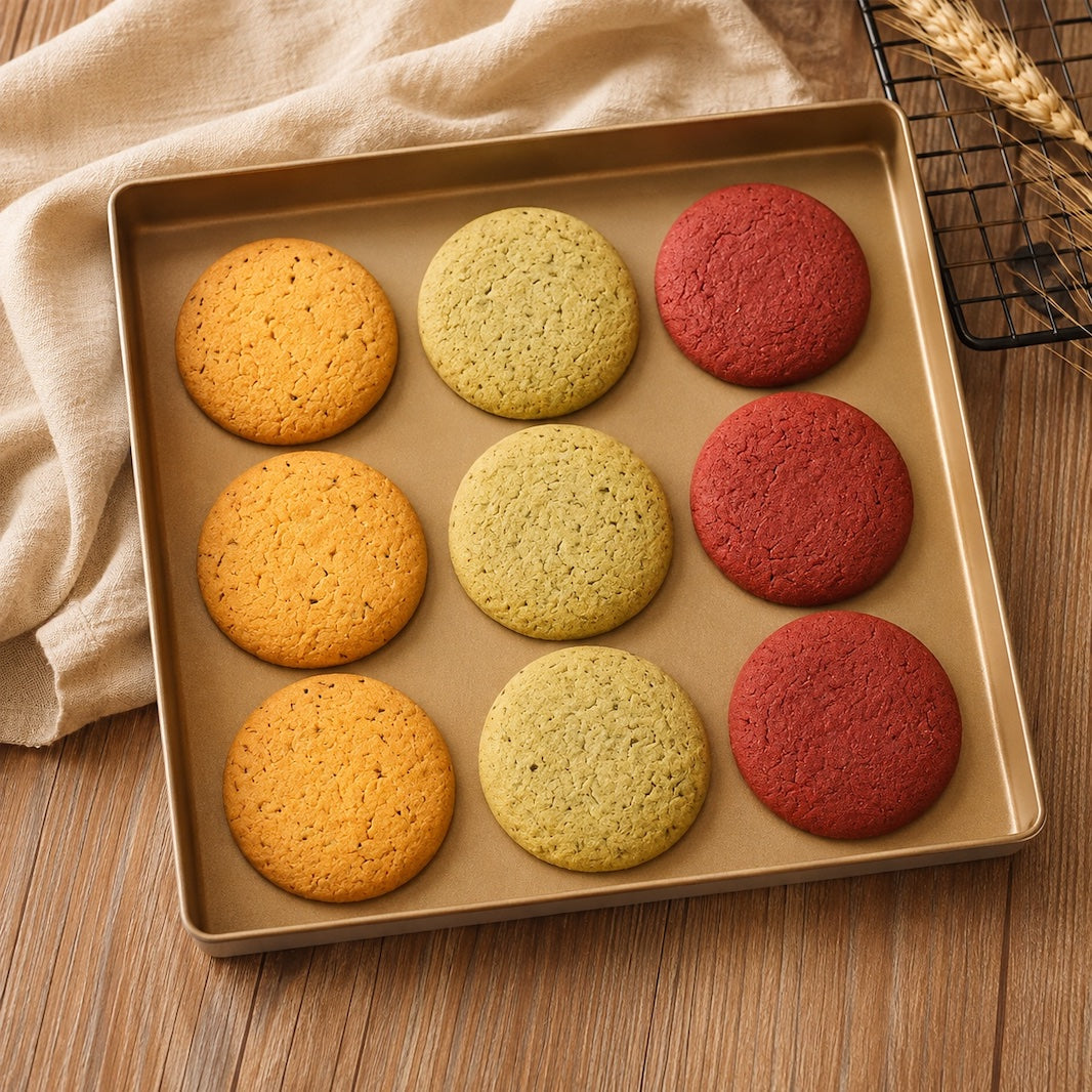 A square Sanneng baking pan holding nine round cookies in three colors: three orange, three yellow-green, and three red. The pan is placed on a wooden table with a beige cloth and wheat stalks in the background.
