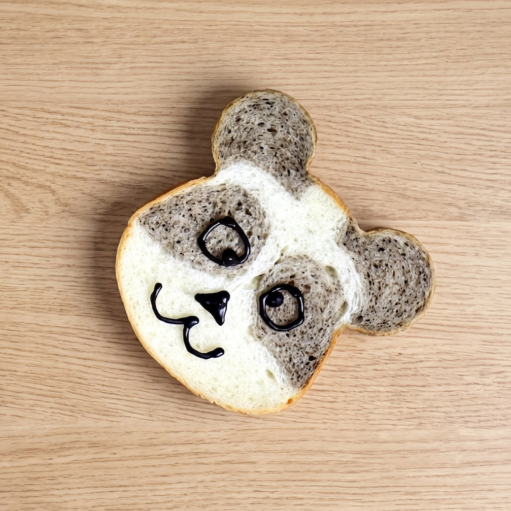 A Sanneng panda-shaped toast made from two slices of bread, with dark chocolate or black icing drawn on to create the panda's face, placed on a light wood grain surface.