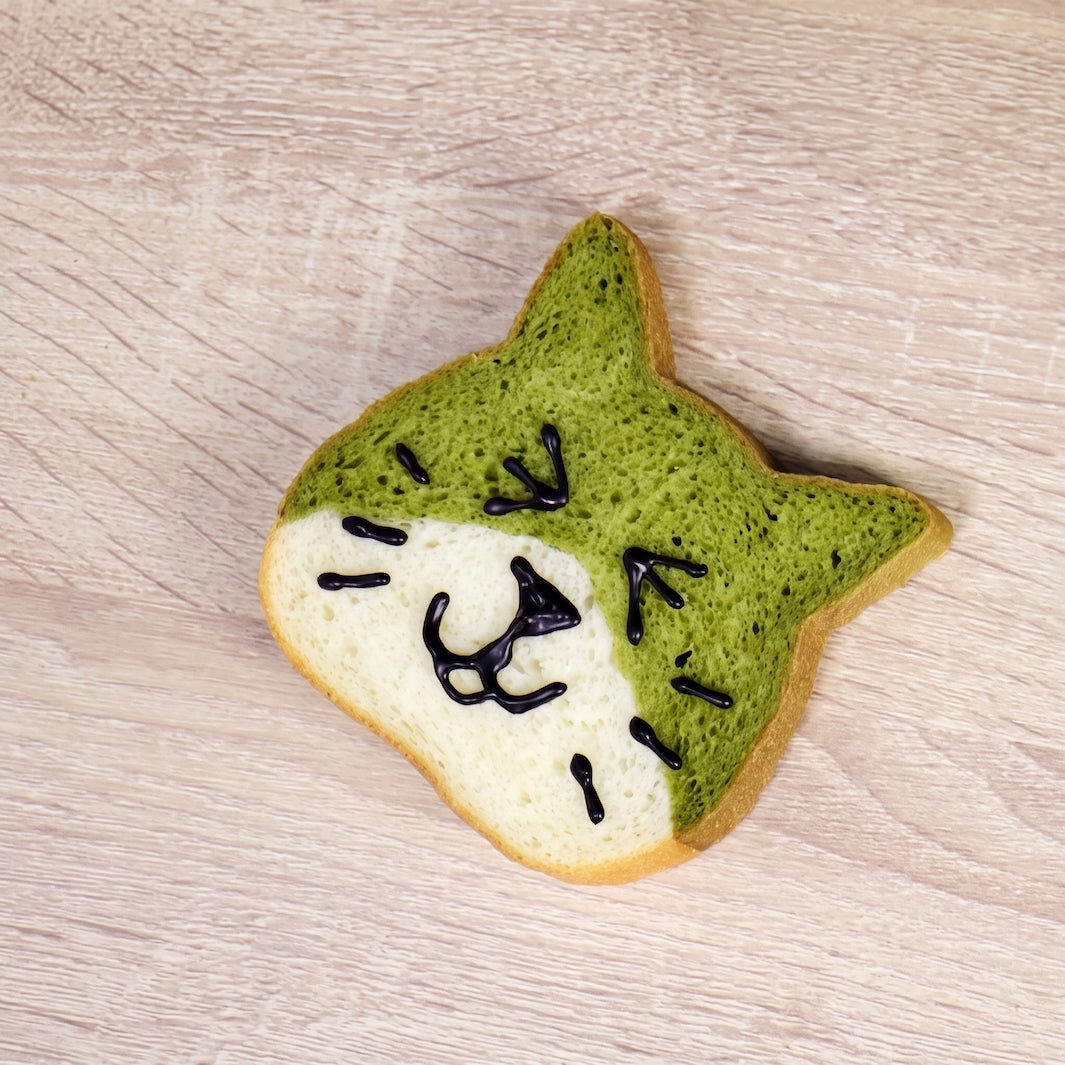 A slice of bread shaped like a cat face, with one half green matcha-style and the other half white, decorated with black icing whisk marks, placed on a light wood surface.