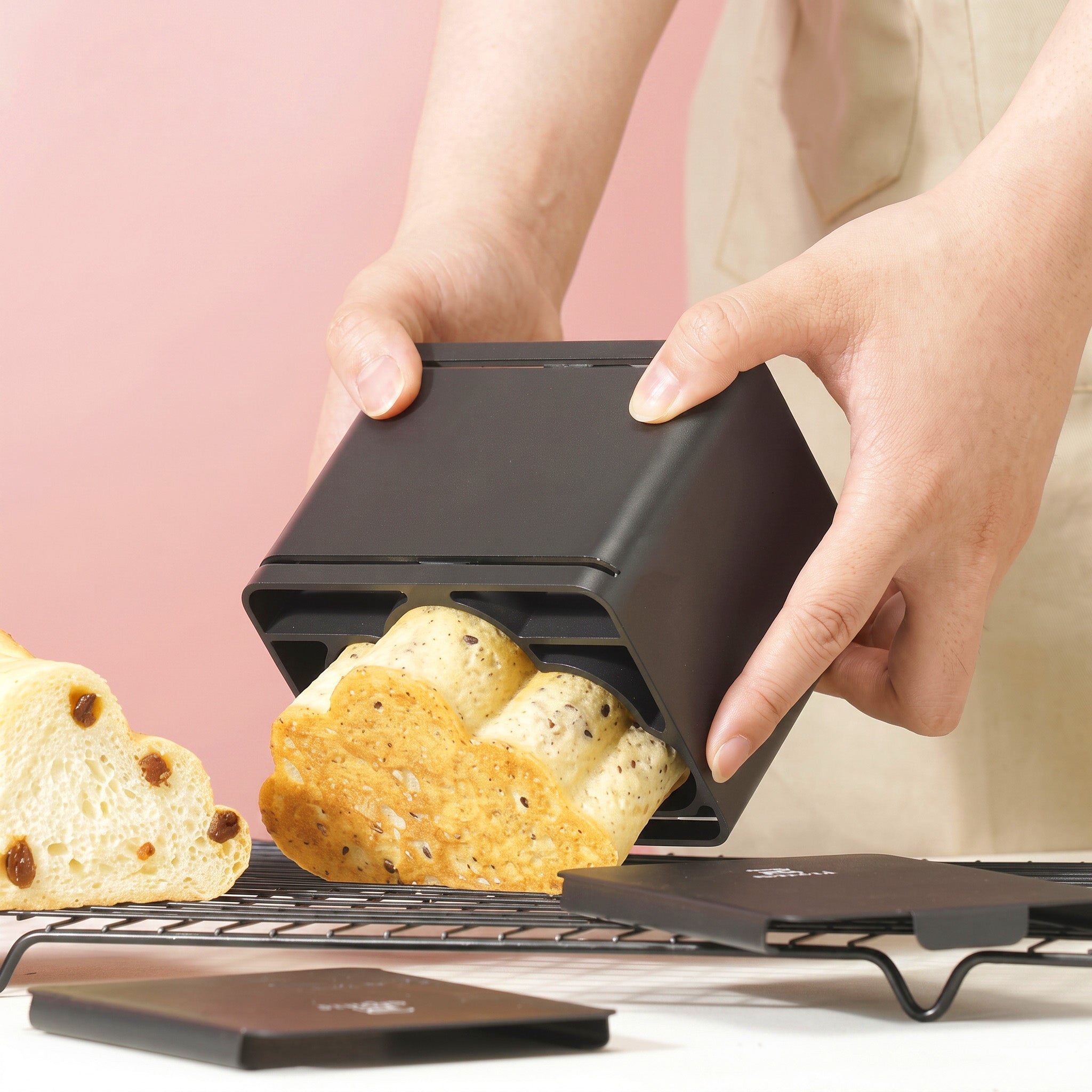 A person's hands are shown using Sanneng black rectangular bread mold to shape a loaf of bread, with the finished loaf sliding out. Another loaf and a lid are visible on a cooling rack against a soft pink background.