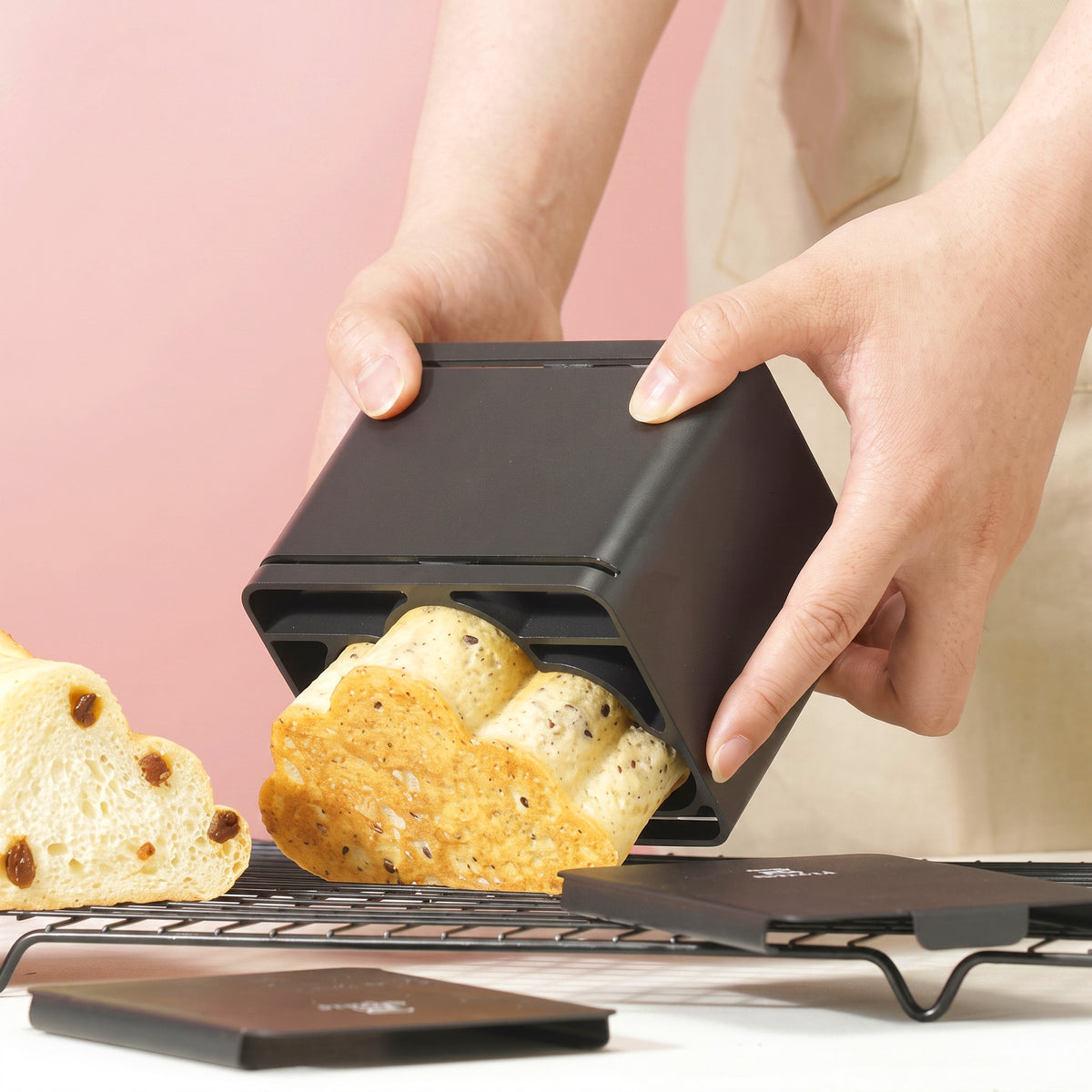A person's hands are shown using Sanneng black rectangular bread mold to shape a loaf of bread, with the finished loaf sliding out. Another loaf and a lid are visible on a cooling rack against a soft pink background.