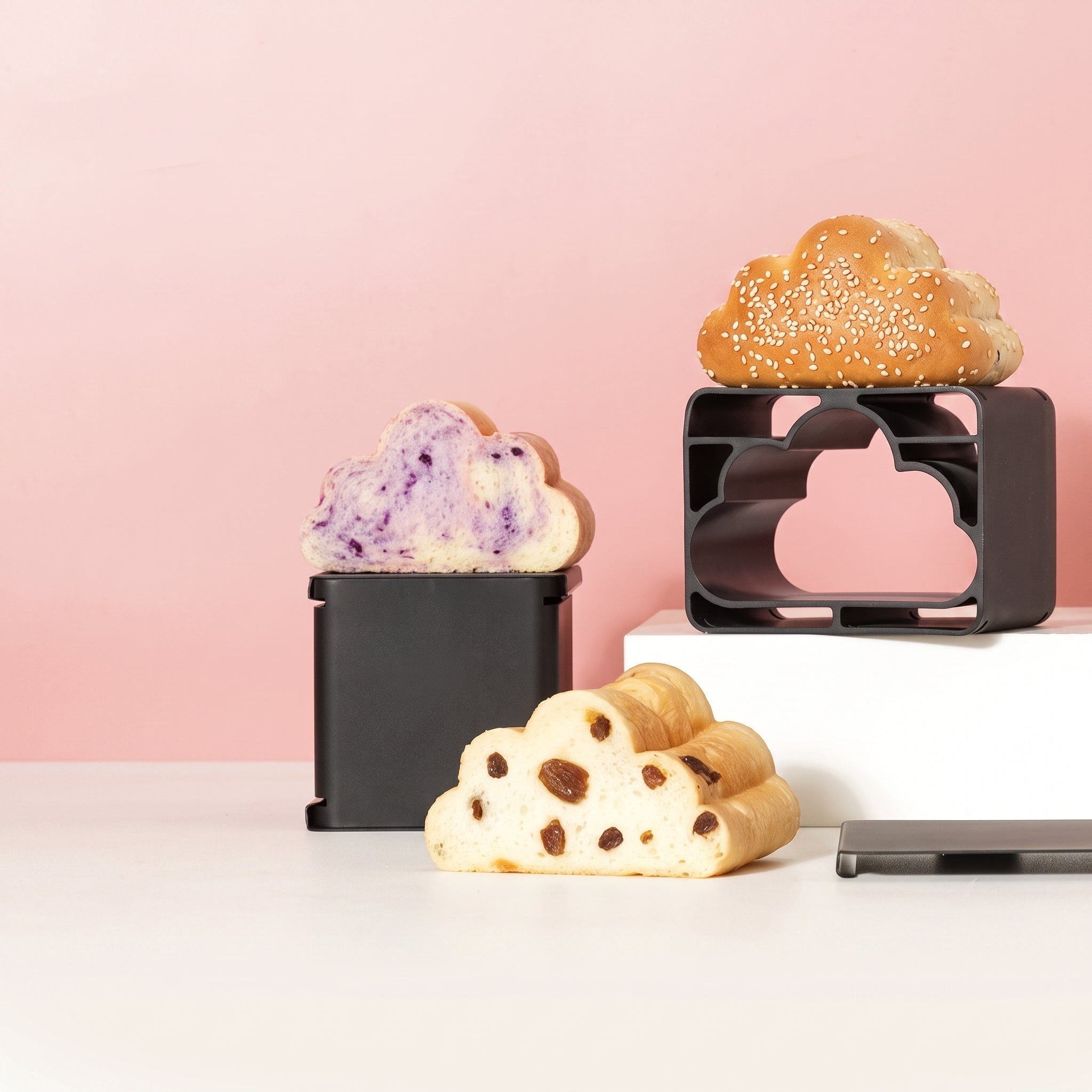 Three cloud-shaped bread loaves displayed on black cloud-shaped loafers against a soft pink background, with one loaf on a white pedestal and two on the tabletop.