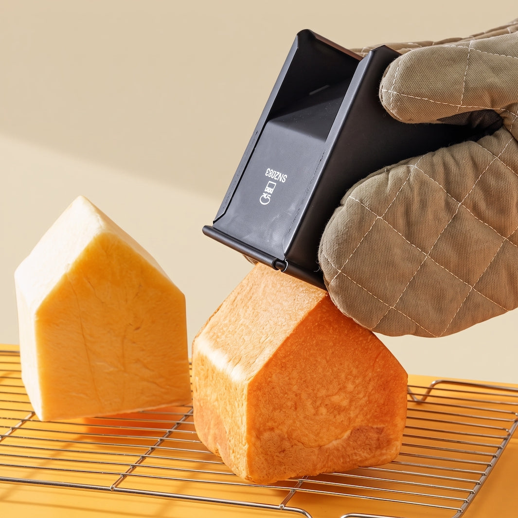 A hand wearing a quilted beige oven mitt holds a black Sanneng shokupan mold over a loaf of bread, with two house-shaped loaves resting on a metal cooling rack against a warm beige background.