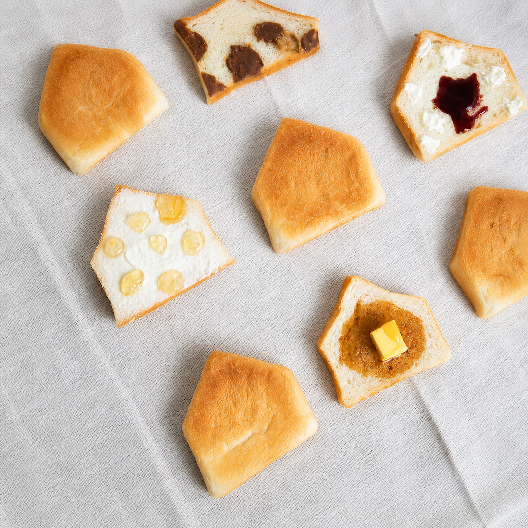 Seven pieces of artisanal white bread, each cut into a pentagonal slice, arranged on a light gray textured fabric background. Some slices are plain, while others are spread with butter, jam, or cheese.