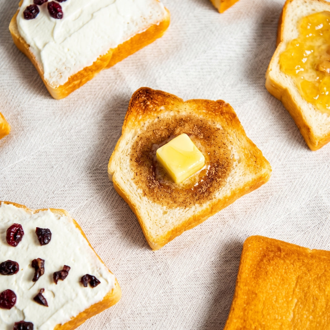 Close-up of toasted bread slices on a light fabric surface, including one with a butter cube in the center and others with cream, jam, or dried fruit toppings.
