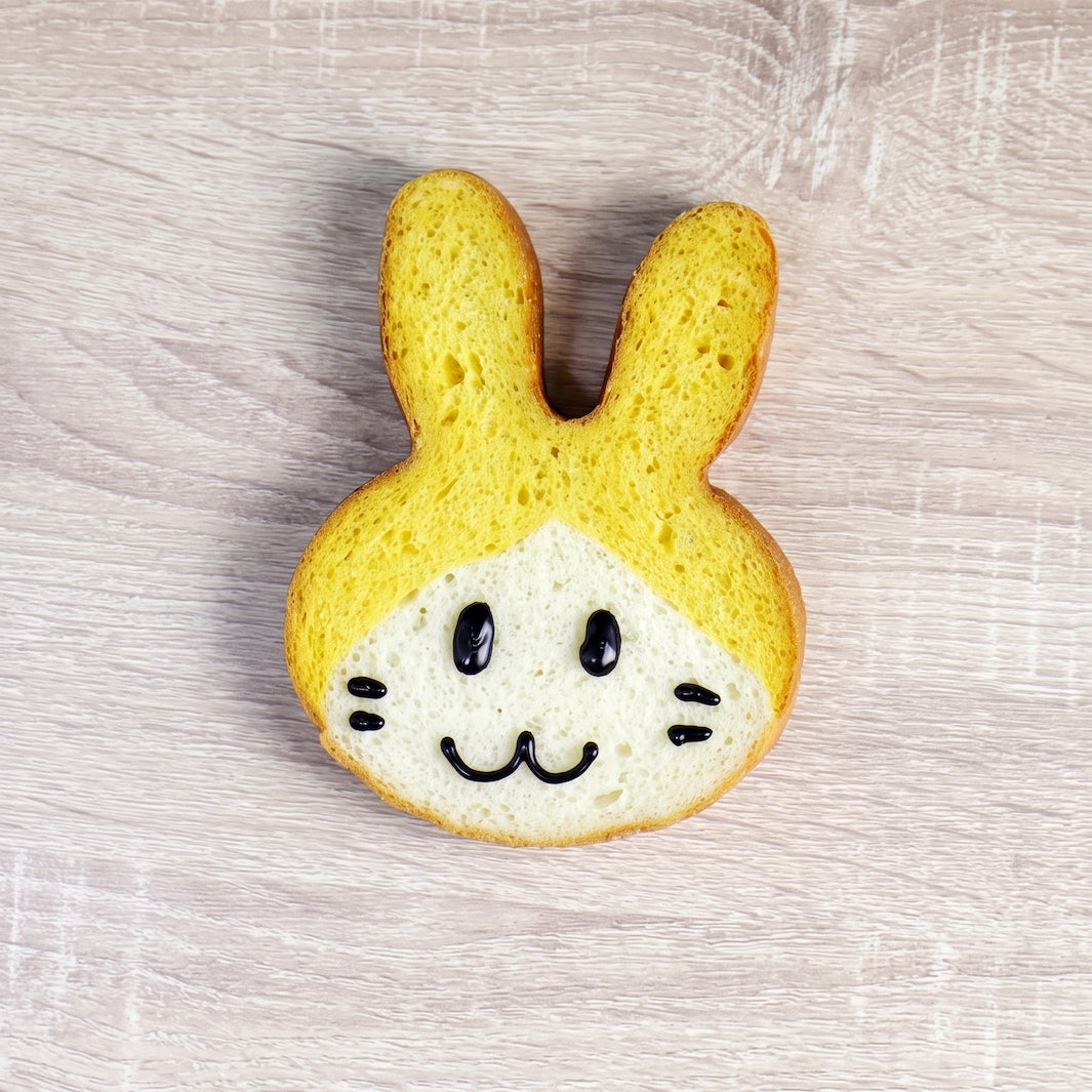 A rabbit-shaped toast made with two slices of bread, featuring yellow cheese-like filling on top and a white bread base with a black icing face, placed on a light wood surface.