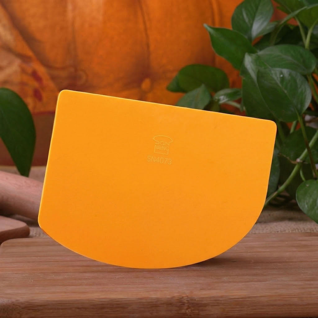 Orange SN4072 Sanneng dough scraper with a brand logo on a wooden surface with a pumpkin and plants in the background