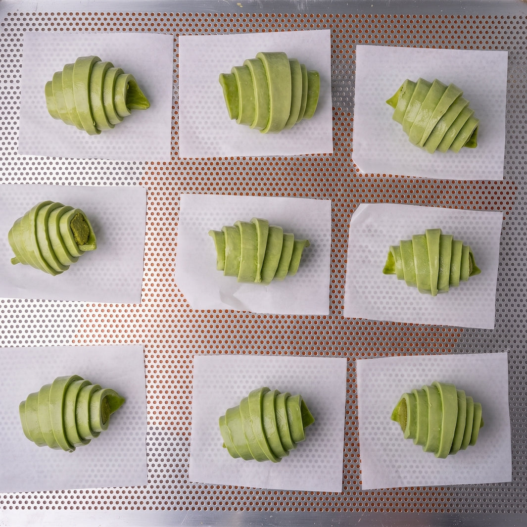 Nine green, spiral-sliced pastry pieces arranged on white parchment paper over a perforated metal baking tray.