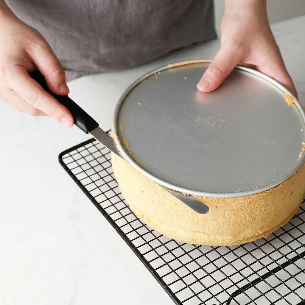 Person using a Sanneng stainless steel cake remover tool with plastic handler to remove a cake on a cooling rack.