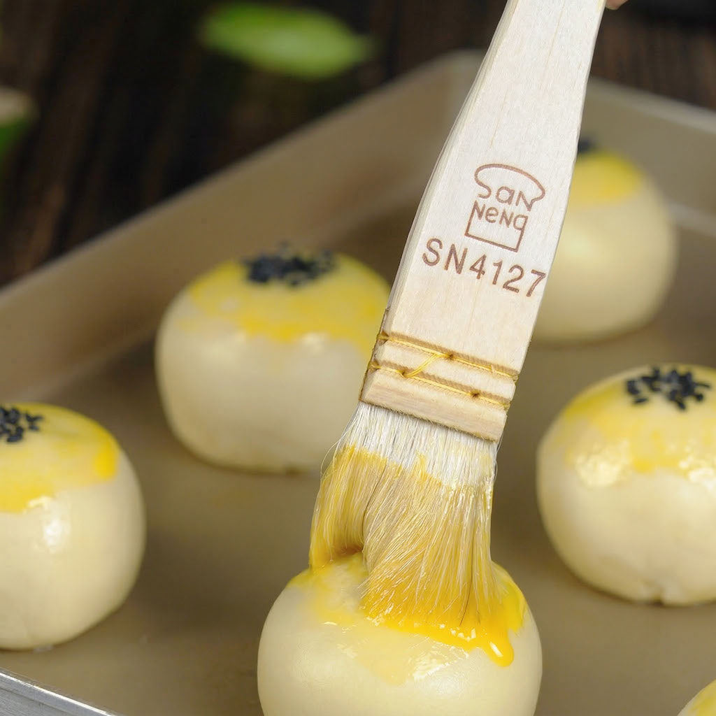 Sanneng pastry brush applying yellow glaze to round baked goods on a tray.