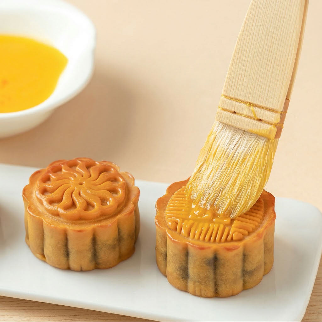 Two mooncakes being brushed by a Sanneng wooden pastry brush with a yellow glaze on a white plate.