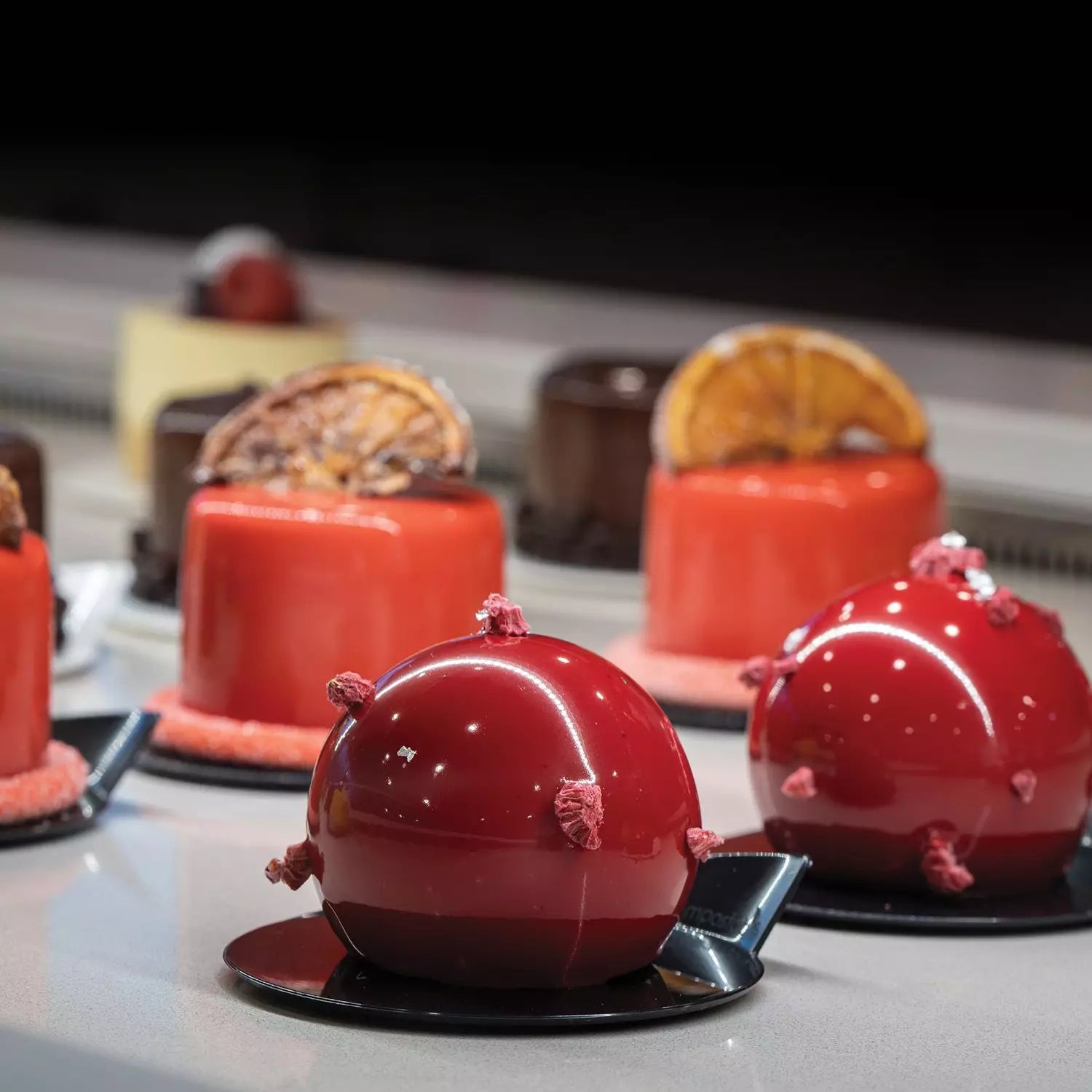 Delicate red dessert with decorative elements on a black plate, surrounded by other pastries.