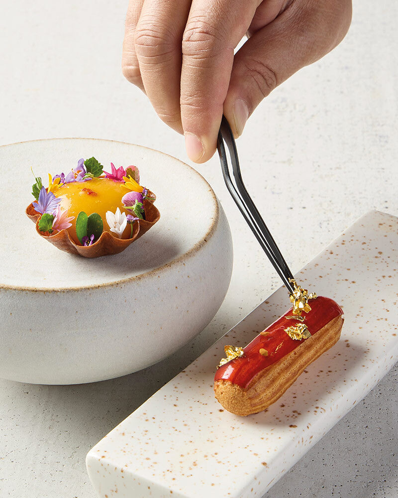 Delicate eclair being garnished with a small flower, with a white bowl and speckled surface in the background.