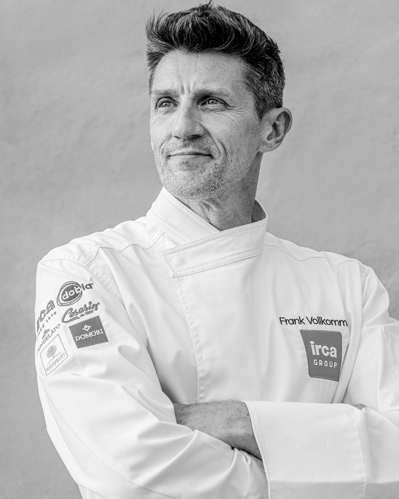 Black and white portrait of chef Frank Vollkomm with visible brand logos on his jacket.