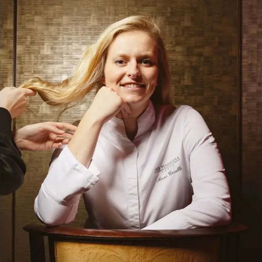 Chef in a white chef's coat sitting in a chair with a person adjusting her hair against a wooden background.
