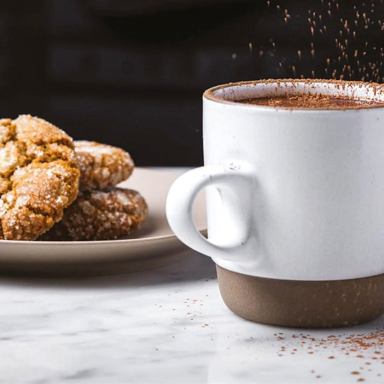 White mug with brown accents filled with a hot chocolate, next to cookies on a marble surface.
