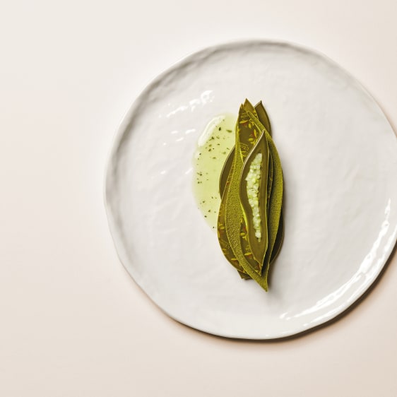 A top-down view of a white textured ceramic plate on a light beige background, featuring a small artistic serving of green dessert made of Valrhona Matcha Inspiration and a drizzle of oil.