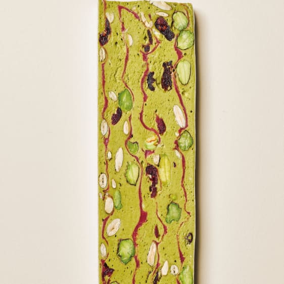 A long rectangular green matcha inspiration chocolate bar with embedded pistachios, almonds, and dried fruit pieces, shown against a plain off-white background.
